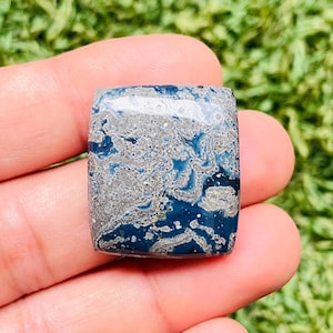 Könnte beinhalten: Ein rechteckiger Edelstein mit einem marmorierten Muster in Tiefblau und Grau. Der Stein wird in einer Hand gehalten, mit einem verschwommenen grünen Hintergrund. Die polierte Oberfläche reflektiert das Licht und hebt die komplizierte Maserung hervor.