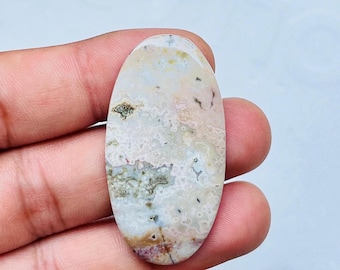 Könnte beinhalten: Ein ovaler Edelstein mit einem gesprenkelten Muster aus Weiß, Beige und Grau. Der Stein hat Bereiche in Hellbraun und Lila, mit kleinen dunklen Punkten. Die polierte Oberfläche reflektiert das Licht und hebt die natürlichen Farb- und Texturvariationen hervor.