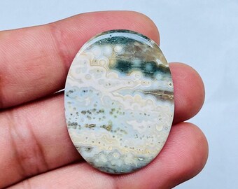 May include: Oval-shaped gemstone with a unique pattern of colors. The stone features a mix of white, beige, green, and brown hues, creating a marbled effect. The polished surface reflects light, highlighting the natural variations in the stone.