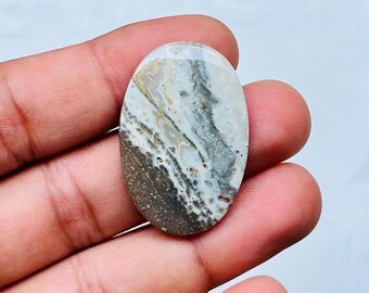 May include: An oval-shaped gemstone with a unique pattern of brown, gray, and white swirls. The polished stone is held in a hand, showcasing its natural colors and textures. The gemstone's surface has a glossy finish.