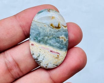 May include: An oval-shaped gemstone with a polished surface. The stone features a blend of colors, including blue, cream, beige, and hints of red. The pattern resembles a landscape with layered textures and variations in color.