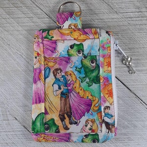 Puede incluir: Una bolsa de tela blanca con cremallera y una ventana de plástico transparente. La bolsa presenta un estampado colorido de una pareja de dibujos animados, una rana y un sol. La bolsa tiene un anillo de metal en la parte superior para sujetar llaves.