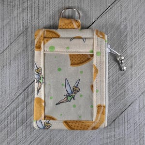 May include: A beige fabric ID badge holder with a clear plastic window. The fabric has a pattern of Tinkerbell fairies and brown cookies. The holder has a zipper and a metal ring.
