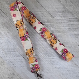 May include: A white lanyard with a repeating pattern of Simba from The Lion King and pink flowers. The lanyard has a silver metal clip.