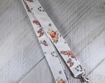 May include: A white fabric lanyard featuring a repeating pattern of Winnie the Pooh, Piglet, and small flower motifs. The strap is constructed from a light-colored textile material and includes a silver-toned metal swivel lobster clasp at the end.