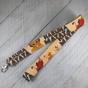 May include: A yellow lanyard with a black and white print of the words "Hakuna Matata" and images of Simba from The Lion King. The lanyard has a black clip on one end.