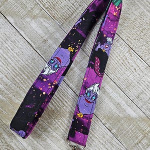 Puede incluir: Un cord&oacute;n morado y negro con una imagen de dibujos animados de &Uacute;rsula de La Sirenita. El cord&oacute;n tiene un cierre plateado y est&aacute; hecho de tela. El fondo del cord&oacute;n tiene un dise&ntilde;o de galaxia.