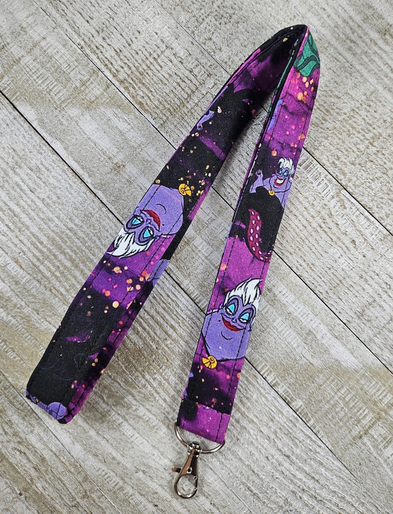Puede incluir: Un cord&oacute;n morado y negro con una imagen de dibujos animados de &Uacute;rsula de La Sirenita. El cord&oacute;n tiene un cierre plateado y est&aacute; hecho de tela. El fondo del cord&oacute;n tiene un dise&ntilde;o de galaxia.