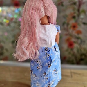 May include: A doll with pink hair and a blue bow. The doll is wearing a white top and a blue skirt with a pattern of snowmen. The doll's hair is long and styled, and the skirt has a button closure.