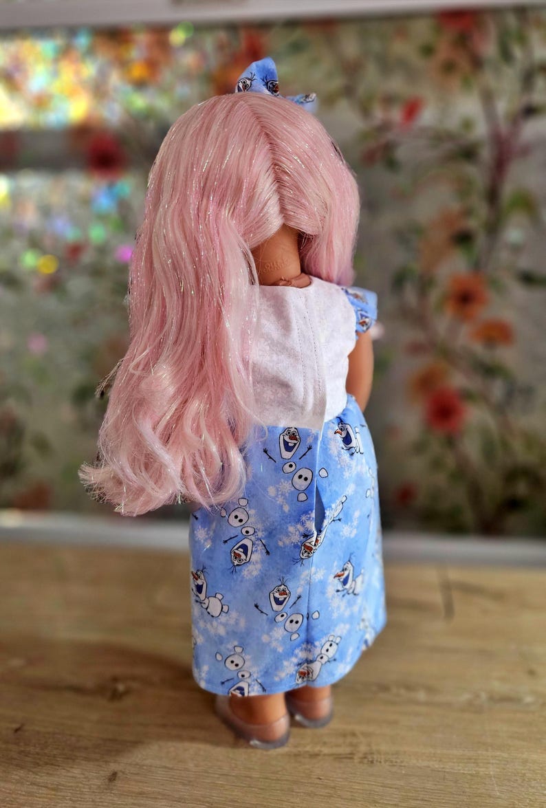 May include: A doll with pink hair and a blue bow. The doll is wearing a white top and a blue skirt with a pattern of snowmen. The doll's hair is long and styled, and the skirt has a button closure.