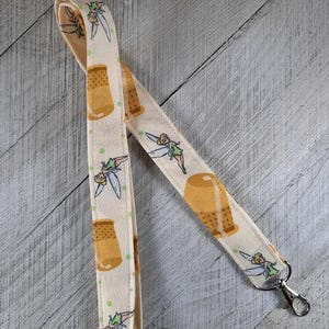 May include: A beige fabric lanyard with a repeating pattern of a cartoon fairy and a thimble. The lanyard has a silver clip on one end.