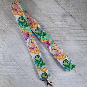 Puede incluir: Un cordón blanco con un patrón repetido de Rapunzel y Pascal el camaleón. El cordón tiene un clip giratorio plateado.