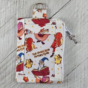 Puede incluir: Cartera de tela blanca con cierre de cremallera y llavero. La cartera presenta un patrón repetido de personajes de dibujos animados y el texto "THE ONCE AND FUTURE KING".