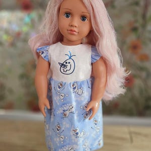 May include: A doll with pink hair, blue eyes, and a blue bow. The doll wears a white and blue dress with a cartoon character design. The dress has short sleeves and a matching bow. The doll's skin is a light peach color.