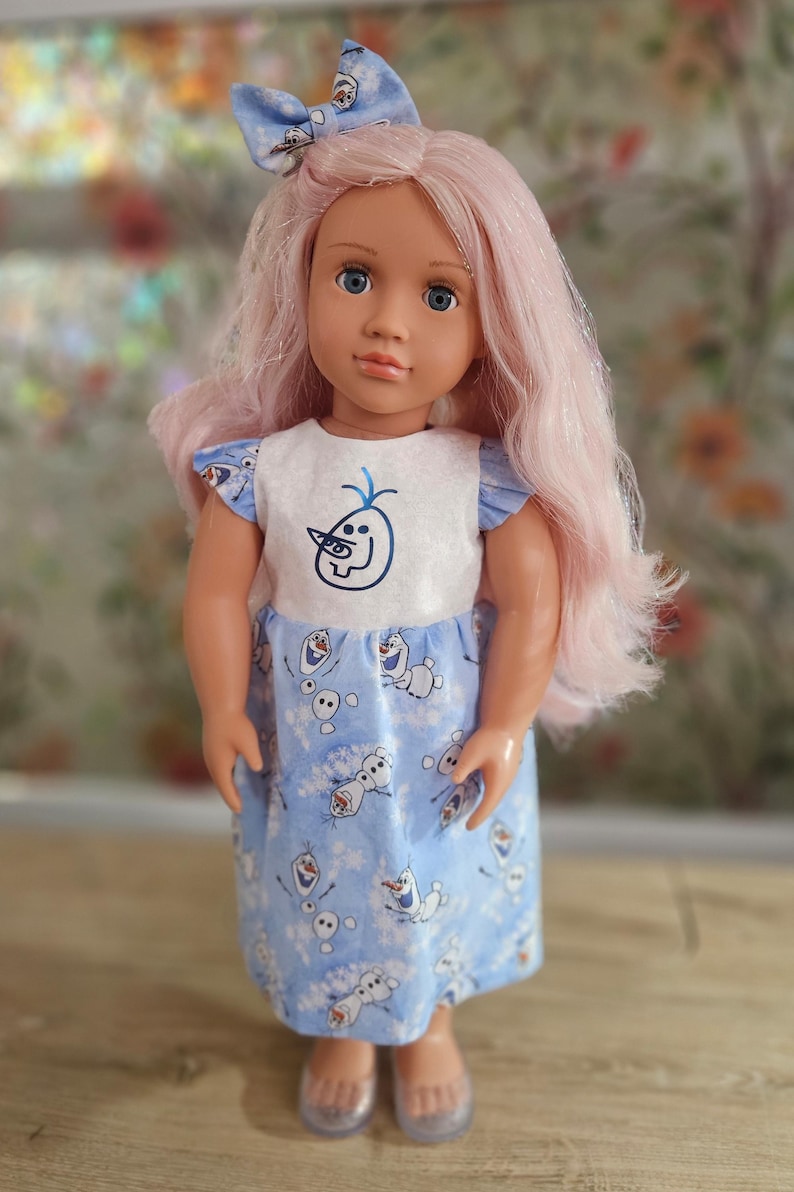 May include: A doll with pink hair, blue eyes, and a blue bow. The doll wears a white and blue dress with a cartoon character design. The dress has short sleeves and a matching bow. The doll's skin is a light peach color.