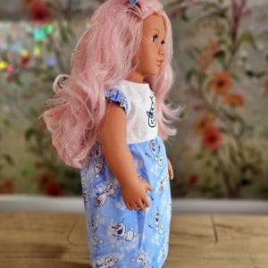 May include: A doll with pink hair and a blue and white dress featuring Olaf from Frozen. The doll has a matching blue bow in its hair and is wearing clear shoes. The dress has a white top with a character design.