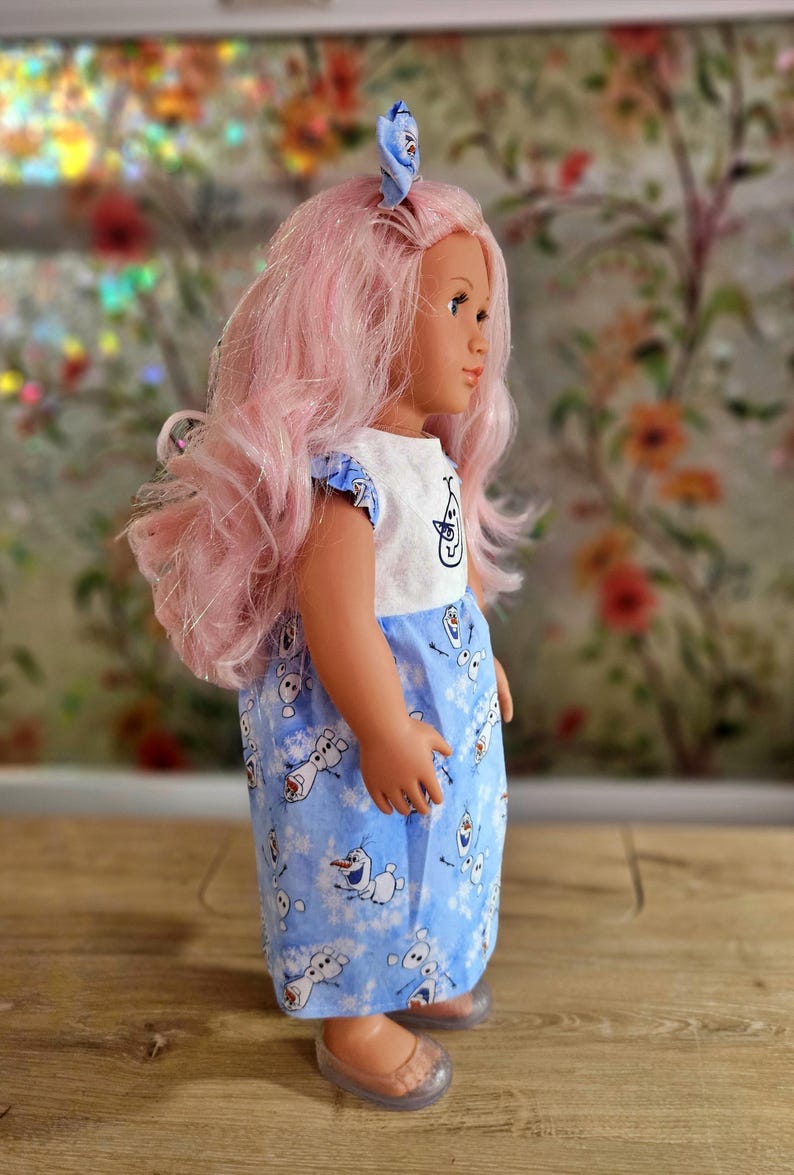 May include: A doll with pink hair and a blue and white dress featuring Olaf from Frozen. The doll has a matching blue bow in its hair and is wearing clear shoes. The dress has a white top with a character design.