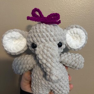 May include: A gray crocheted elephant plush toy with white ears and black eyes. The elephant is wearing a purple bow on its head.