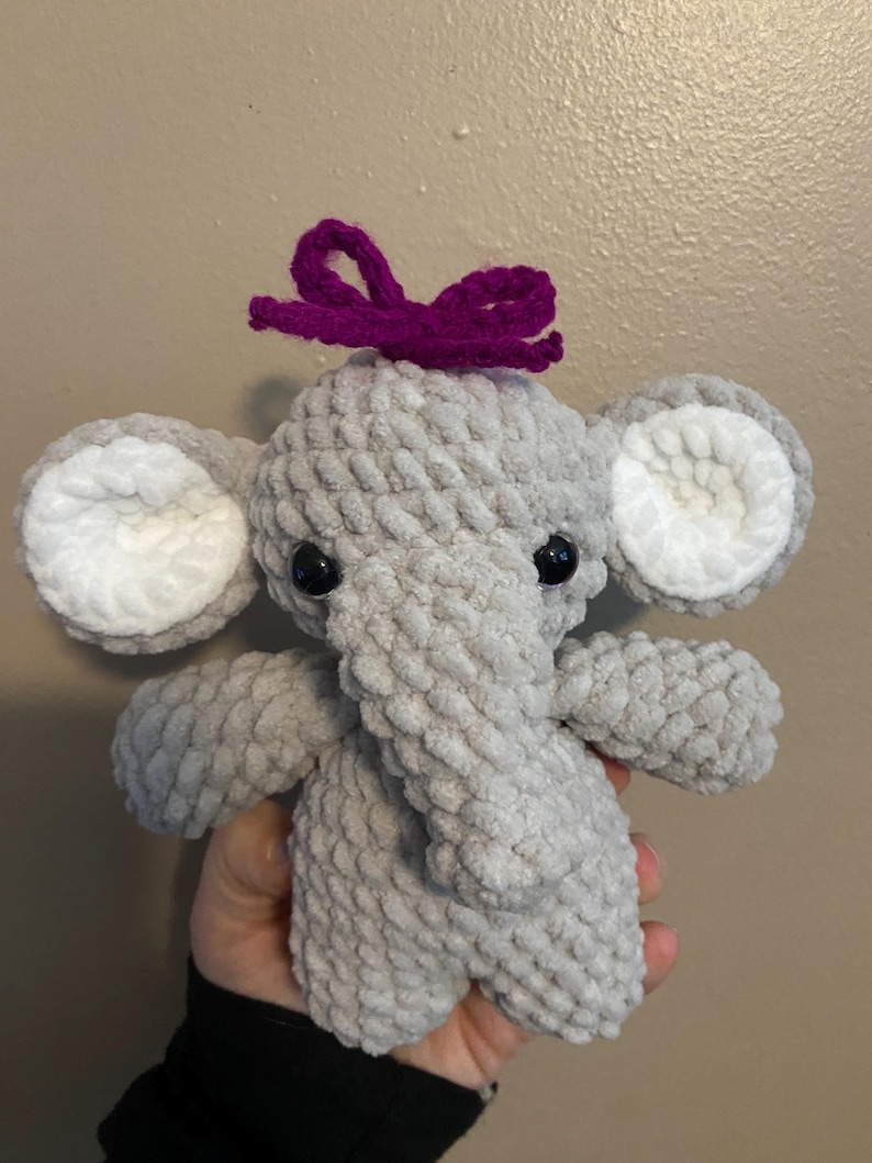 May include: A gray crocheted elephant plush toy with white ears and black eyes. The elephant is wearing a purple bow on its head.