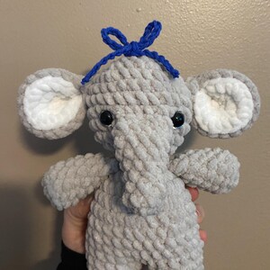 May include: A gray crocheted elephant plush toy with white ears and a blue bow on its head.