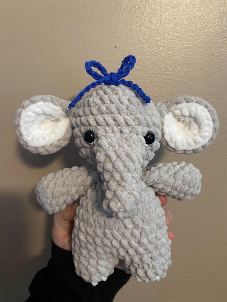 May include: A gray crocheted elephant plush toy with white ears and a blue bow on its head.
