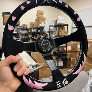 May include: Black steering wheel with pink cherry blossom accents and Japanese characters. The wheel has a black center and spokes, with white stitching and the text "Doll Boost Performance".