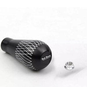 Peut inclure: Un pommeau de levier de vitesses noir avec un motif en losanges et le texte "Bull Boost PERFORMANCE" en blanc. Un écrou argenté est à côté du pommeau. Le pommeau est conçu pour une utilisation automobile.