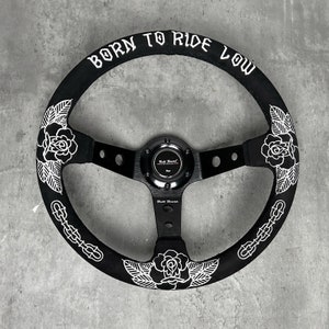 May include: Black steering wheel with white floral and chain designs. The top of the wheel has the text "BORN TO RIDE LOW" in white. The center of the wheel has a black cap with the text "Built Boost".