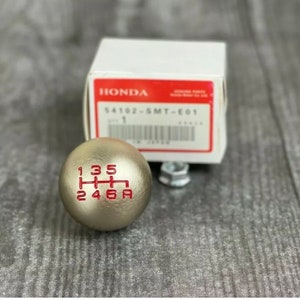 May include: A silver shift knob with red gear markings, including the numbers 1, 2, 3, 4, 5, and 6, and the letter A. The knob is next to a white box with the Honda logo and product information.