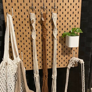 May include: A wooden pegboard displaying macrame plant hangers and crocheted bags. The hangers are cream and brown, and the bags are cream and tan. A small white planter with a green plant is also attached to the board.