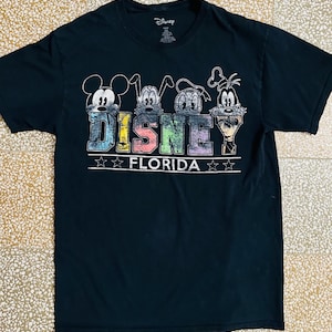 Peut inclure: T-shirt noir avec un motif vintage de Mickey Mouse, Donald Duck, Dingo et Pluto. Le motif comprend le texte "DISNEY FLORIDA" avec des étoiles.