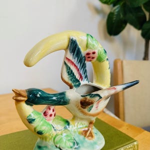 May include: A ceramic duck planter featuring a yellow crescent moon design. The duck is painted in shades of green, brown, and red with a white belly. The planter has a small opening for planting.