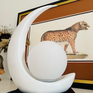 Peut inclure: Lampe en céramique blanche avec un design de croissant de lune tenant une lumière sphérique. La lampe est posée sur un livre noir intitulé "ONE-WAY TICKET". Un imprimé léopard encadré est en arrière-plan.