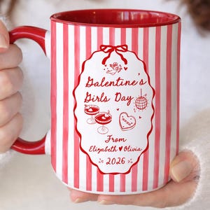 May include: A white ceramic mug with red vertical stripes and a red interior. The mug features a decorative oval with the text "Galentine's Girls Day" and illustrations of cocktails, a heart-shaped cookie, and a disco ball.