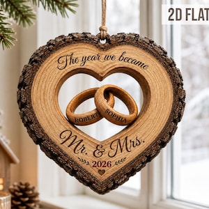 Puede incluir: Un adorno de madera en forma de corazón con bordes de corteza, que presenta el texto "The year we became Mr. & Mrs. 2026" y dos anillos de madera entrelazados grabados con los nombres "Robert" y "Sophia" suspendidos en el centro por un cordel.