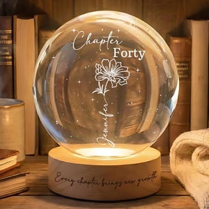 May include: A clear glass sphere with the words "Chapter Forty" and "Jennifer" etched on it, along with a floral design. The sphere sits on a wooden base with the inscription "Every chapter brings new growth." The sphere is illuminated, creating a warm glow.