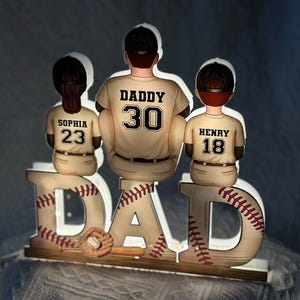 May include: A white wooden sign with the word "DAD" in large letters. Each letter is decorated with a baseball design. Three figures of baseball players stand on top of the sign. The figures are wearing baseball uniforms with the names "Sophia", "Daddy", and "Henry" and the numbers "23", "30", and "18" on the back.