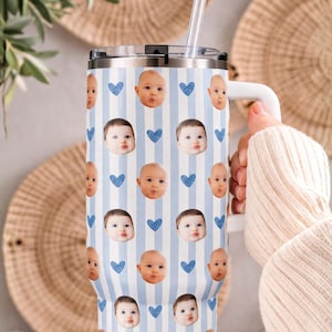 May include: A stainless steel tumbler with a white handle and a clear straw. The tumbler features a pattern of baby faces, blue hearts, and vertical blue and white stripes. The tumbler is designed to keep beverages cold or hot.