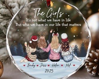 May include: A clear, round Christmas ornament with a red ribbon. The ornament features an illustration of four friends in winter attire, with text that reads "The Girls." Below the illustration are the names Becky, Jess, Helen, and Mel, with the year 2025.