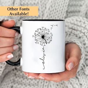 May include: White ceramic mug with a black handle. The mug has a black rim and features a black and white illustration of a dandelion with the word "olivia" written vertically below it.