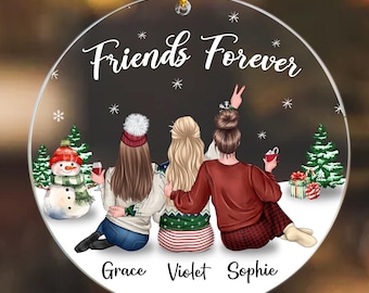 May include: A round Christmas ornament with a white background and the text "Friends Forever" in black. Three cartoon women are sitting in front of a snowman and a Christmas tree. The women are wearing winter clothes and holding glasses of wine. The names "Grace", "Violet", and "Sophie" are written below the women.