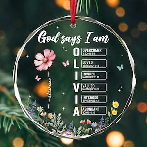 Könnte beinhalten: Ein klarer, runder Weihnachtsschmuck mit dem Text "God says I am" und dem Namen "Olivia". Er zeigt eine rosa Blume, Schmetterlinge und florale Akzente. Der Text listet Eigenschaften wie "Überwinder" und "Geliebt" auf.