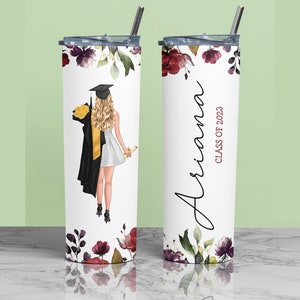 May include: A white stainless steel tumbler with a floral design and a graduation cap illustration. The tumbler has a black lid and a straw. The text "Ariana Class of 2023" is printed on the tumbler.