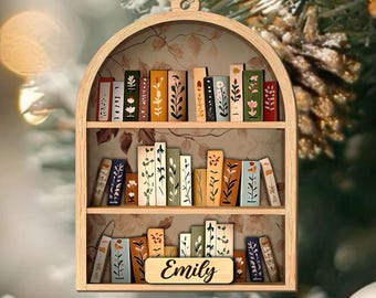 May include: Wooden bookshelf ornament with a decorative arch design. The ornament features three shelves filled with miniature books with floral and leaf designs. The name "Emily" is displayed on a small wooden plaque.