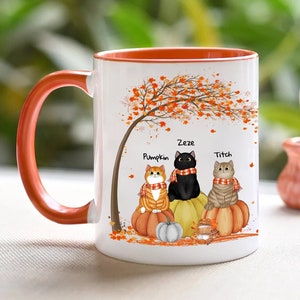 May include: White ceramic coffee mug with an orange rim. The mug features a fall-themed design with three cats sitting under a tree with orange, yellow, and red leaves. The cats are wearing scarves and are named Pumpkin, Zeze, and Titch.