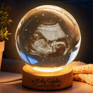 Pode incluir: Uma esfera de vidro transparente com uma imagem de ultrassom no interior, repousando sobre uma base de madeira iluminada. A base tem a inscrição "Baby Sophia ARRIVING 2026". A esfera é decorada com estrelas.