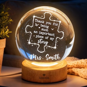 Può includere: Una sfera di vetro trasparente con un design a forma di pezzo di puzzle, illuminata da una luce calda. Il testo recita: "Grazie per essere un pezzo importante della mia storia, Sig.ra Smith." La sfera poggia su una base di legno con il nome "Amanda" inciso.