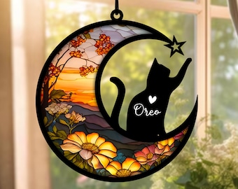 Può includere: Un acchiappasole in vetro colorato a forma di luna con una silhouette nera di un gatto che raggiunge una stella. Il gatto ha un cuore con il nome "Oreo" scritto al suo interno. La luna ha un design in vetro colorato con fiori e un tramonto.