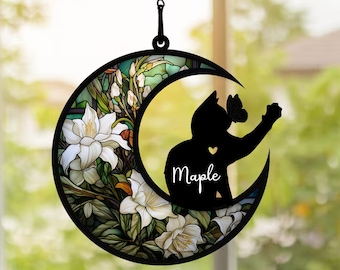 Può includere: Un acchiappasole a forma di mezzaluna in metallo nero con un design in vetro colorato che presenta fiori bianchi e una silhouette di un gatto con la parola "Maple" sul petto.