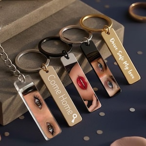 Op de afbeelding: Een verzameling sleutelhangers met rechthoekige hangers. Sommige hangers tonen afbeeldingen van ogen en lippen, terwijl andere de tekst "Come Home" en "Drive Safe My Love" hebben. De sleutelhangers hebben zilveren, zwarte en gouden ringen.